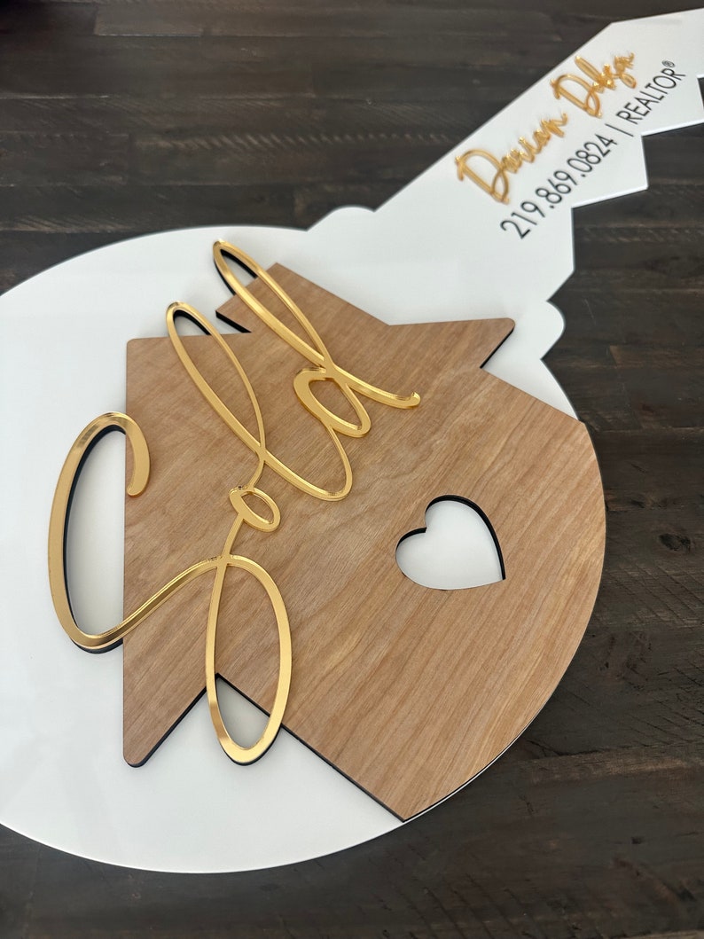 May include: A white and wood house-shaped key with the word "Sold" in gold lettering. The key has a black outline and a small heart cutout. The key is attached to a white and gold key with the text "Davin Diga 219.869.0824 | REALTOR" in black lettering.