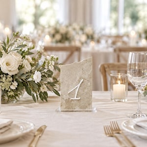 May include: A rectangular beige stone table number sign with a rough, uneven top edge and a white handwritten numeral 1 on the front. The stone slab is supported by a clear acrylic base, standing upright on a light-colored fabric tablecloth.