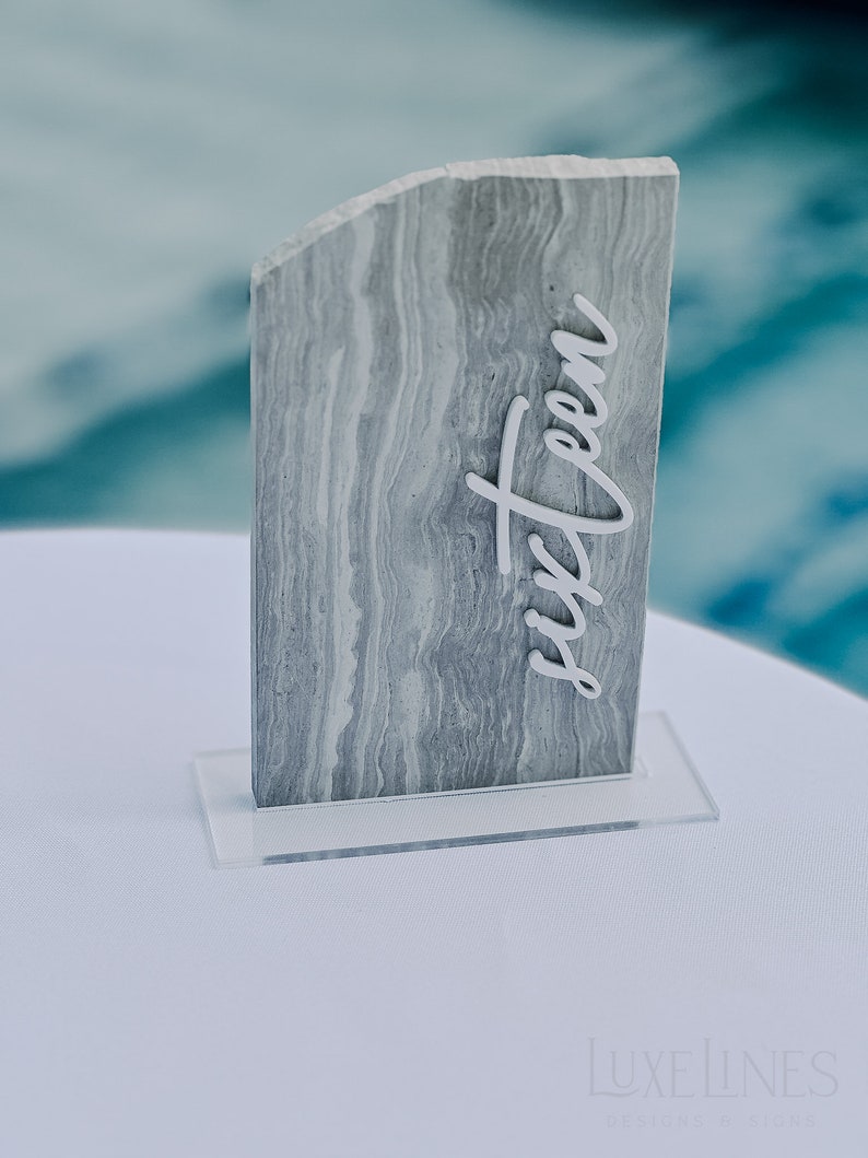 Op de afbeelding: Een grijs en wit gemarmerd tafelnummer zestien, met een heldere acryl basis. Het nummer staat in een cursief lettertype en is bevestigd aan een rechthoekige steen. Het tafelnummer staat op een wit oppervlak.