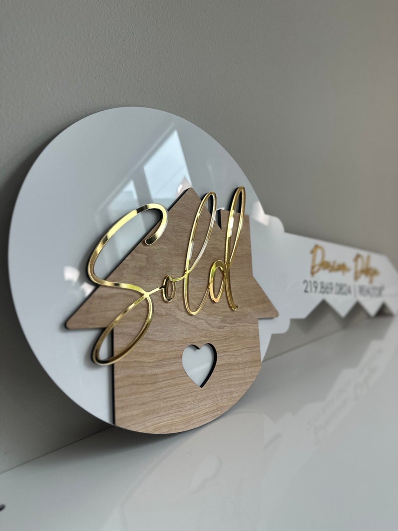 May include: A white and wood house-shaped key with a gold "Sold" sign on top. The key has a heart cut out of the wood and a white background with a realtor's name and phone number.