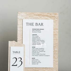 May include: A bar menu and table number 23 are displayed on a beige stone-like background. The bar menu lists signature drinks, liquor, wine, and beer. The table number is in a simple black font.
