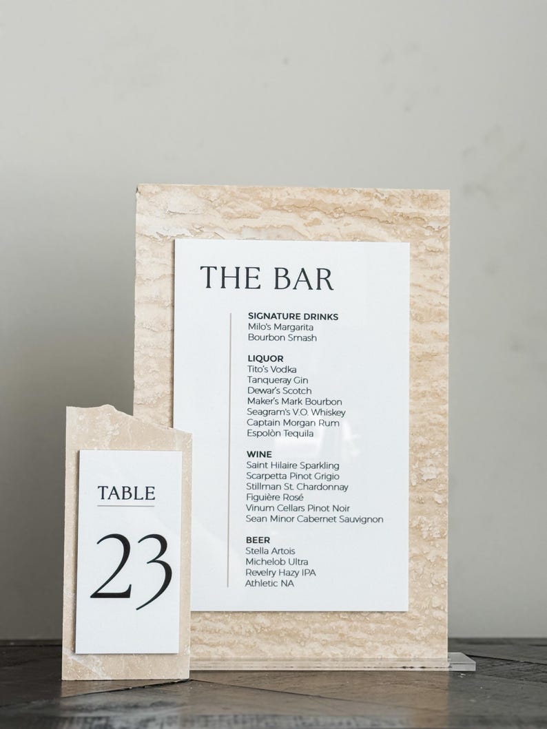 May include: A bar menu and table number 23 are displayed on a beige stone-like background. The bar menu lists signature drinks, liquor, wine, and beer. The table number is in a simple black font.
