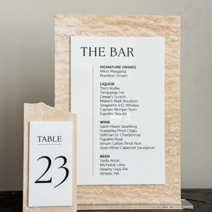 May include: A bar menu and table number 23 are displayed. The bar menu lists signature drinks, liquor, wine, and beer options. The table number is on a separate card. Both cards have a textured, beige background.