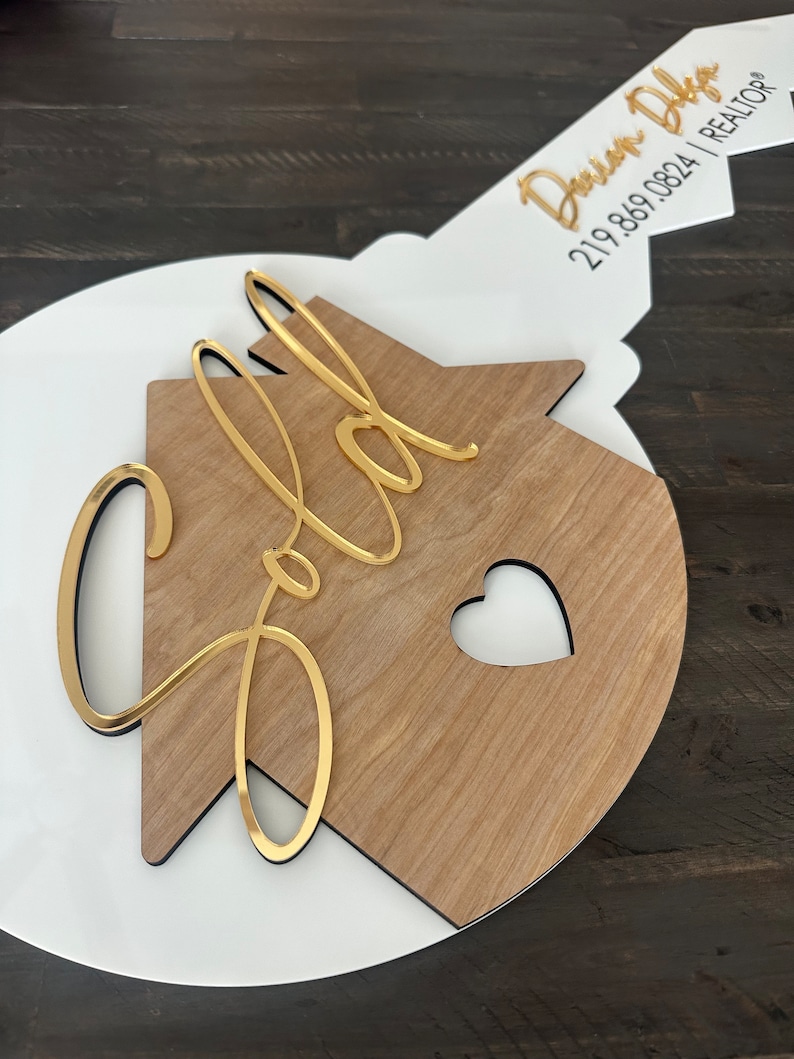 May include: A white and wood house-shaped key with a gold inscription that reads "Sold" and a heart cutout. The key has a gold tag with the text "Devin Diga 219.869.0824 | REALTOR&reg;".