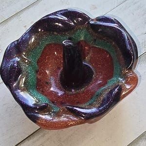 May include: A resin ring holder with a multi-colored glitter design. The holder features a dark purple outer edge, transitioning to green, red, and orange hues. A black post rises from the center. The holder sits on a white wooden surface.