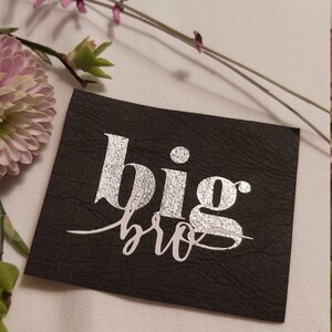 Könnte beinhalten: Ein schwarzer Leder-Patch mit silbernem Glitzer-Schriftzug, der "big bro" lautet.