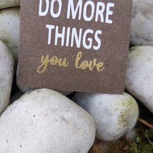 Könnte beinhalten: Eine braune Karte mit weißem Text, der "DO MORE THINGS you love" lautet, auf einem Hintergrund aus weißen Steinen.