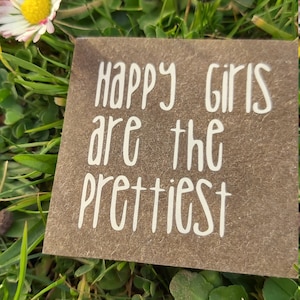 Könnte beinhalten: Eine braune Karte mit weißem Text, der "Happy girls are the prettiest" lautet, auf einem Hintergrund aus grünem Gras und weißen Gänseblümchen.