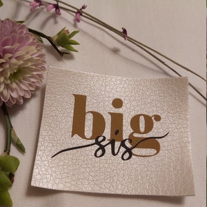 Könnte beinhalten: Ein weißer Kunstleder-Patch mit den Worten "big sis" in goldenen und schwarzen Buchstaben.