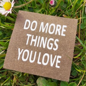 Könnte beinhalten: Eine braune Papierkarte mit dem Text "Do more things you love" in weißen Buchstaben. Die Karte liegt auf einem Bett aus grünem Gras mit weißen Gänseblümchen.
