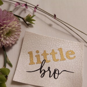 Könnte beinhalten: Weißer Leder-Patch mit den Worten "little bro" in goldenen und schwarzen Buchstaben.