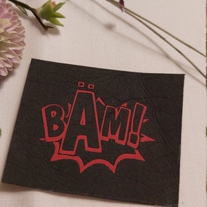 Könnte beinhalten: Schwarzer Leder-Patch mit einer roten "BAM!" Grafik in Comic-Schrift.