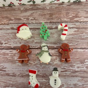 May include: A collection of Christmas-themed decorations, including a Santa Claus, Christmas tree, candy cane, gingerbread people, penguin, stocking, and snowman. The decorations are in various colors and placed on a wooden surface.
