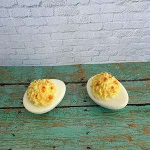 May include: Two deviled eggs on a teal wooden surface against a white brick wall. The eggs are cut in half, with a white egg white and a yellow filling sprinkled with paprika. The eggs are a classic appetizer.