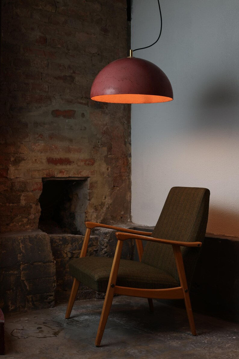 Pu&ograve; includere: Una poltrona marrone con gambe in legno si trova in una stanza con un muro di mattoni e un camino. Un lampadario a cupola rosa &egrave; appeso al soffitto.
