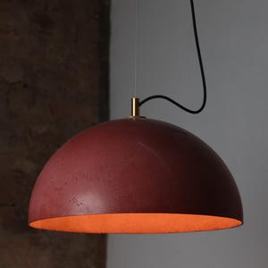 Pu&ograve; includere: Una lampada a sospensione in cemento rosso con una parte superiore in metallo dorato e un cavo nero. La luce &egrave; appesa al soffitto e illumina la stanza.