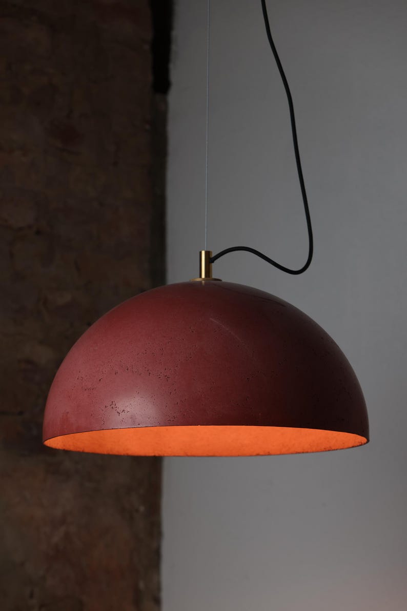 Pu&ograve; includere: Una lampada a sospensione in cemento rosso con una parte superiore in metallo dorato e un cavo nero. La luce &egrave; appesa al soffitto e illumina la stanza.