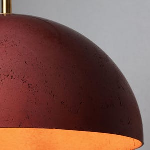 Pu&ograve; includere: Una lampada a sospensione in cemento rosso con finitura dorata. La luce &egrave; accesa ed emette una luce calda.