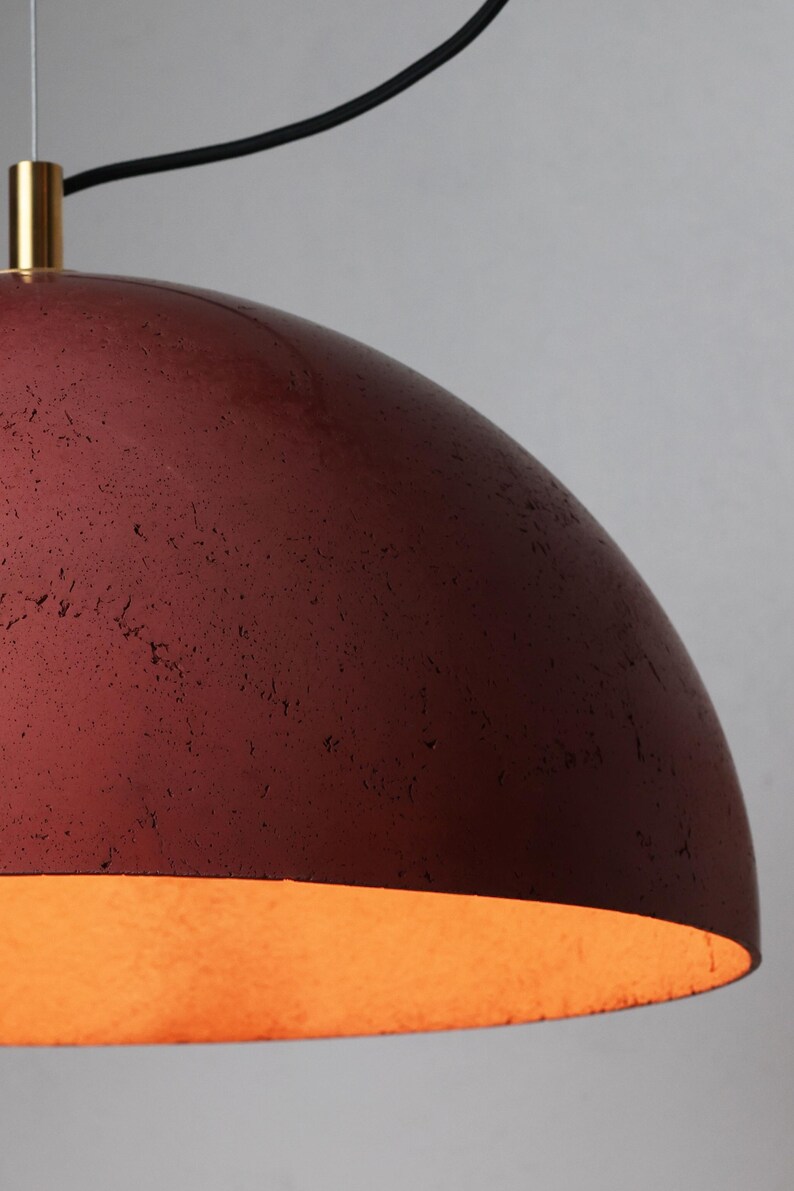 Pu&ograve; includere: Una lampada a sospensione in cemento rosso con finitura dorata. La luce &egrave; accesa ed emette una luce calda.