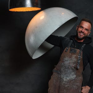 Pu&ograve; includere: Un uomo con un grembiule di cuoio marrone tiene in mano un grande lampadario rotondo in cemento. Altri due lampadari sono appesi sullo sfondo, uno nero e uno arancione.