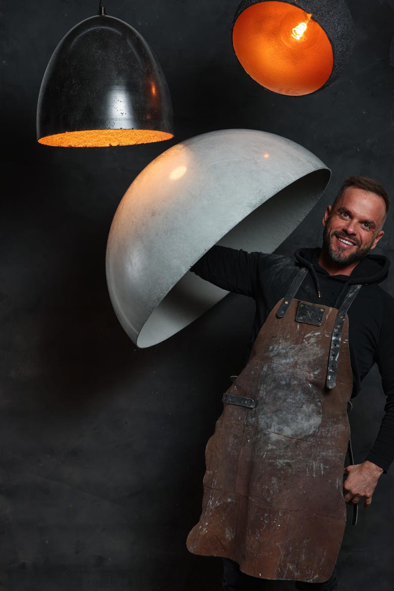 Pu&ograve; includere: Un uomo con un grembiule di cuoio marrone tiene in mano un grande lampadario rotondo in cemento. Altri due lampadari sono appesi sullo sfondo, uno nero e uno arancione.