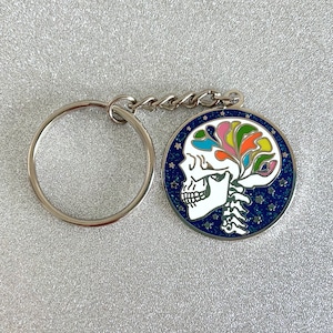 May include: A silver key chain with a round, blue enamel charm featuring a white skull with colorful flowers and stars.