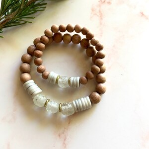May include: A set of two bracelets, one made of brown wooden beads and the other with white and clear glass beads with gold accents.