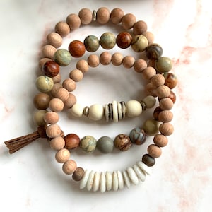 May include: Set of three bracelets featuring natural wood beads, white shell beads, and brown, green, and white patterned beads. The bracelets are stacked on top of each other and have a brown tassel.