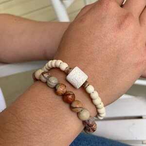 May include: Two bracelets on a wrist. One bracelet is made of white beads with a square white lava rock bead. The other bracelet is made of brown and white patterned beads.
