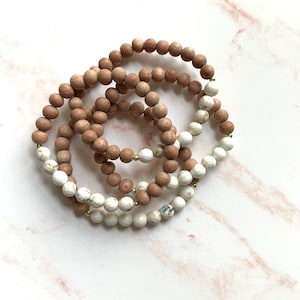 May include: A set of four bracelets featuring brown wooden beads and white and gray stone beads. The bracelets are stacked on top of each other.