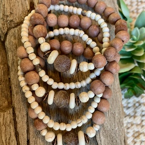 May include: A stack of wooden bead bracelets in shades of brown and cream. The bracelets feature various sizes and shapes of beads, including round and oblong designs. The bracelets are displayed on a piece of weathered wood.
