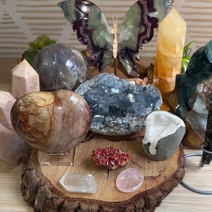 May include: A collection of crystals and stones arranged on a wooden slice. The collection includes a heart-shaped stone, a large blue crystal, a clear crystal, a pink crystal, a red crystal, a clear crystal sphere, a yellow crystal, and a green and purple butterfly sculpture.