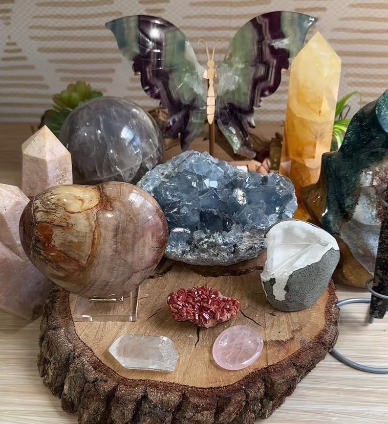 May include: A collection of crystals and stones arranged on a wooden slice. The collection includes a heart-shaped stone, a large blue crystal, a clear crystal, a pink crystal, a red crystal, a clear crystal sphere, a yellow crystal, and a green and purple butterfly sculpture.