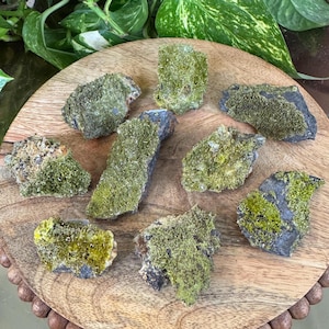 May include: A collection of raw, natural stones with a mossy green and yellow-green texture. The stones are arranged on a round, wooden surface, showcasing their unique shapes and colors. The stones vary in size and shape, with some appearing more rectangular and others more irregular.