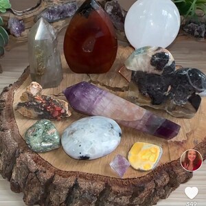 May include: A collection of crystals and stones on a wooden slice. The crystals include amethyst, clear quartz, carnelian, and a white selenite plate. The stones are green, brown, and black.