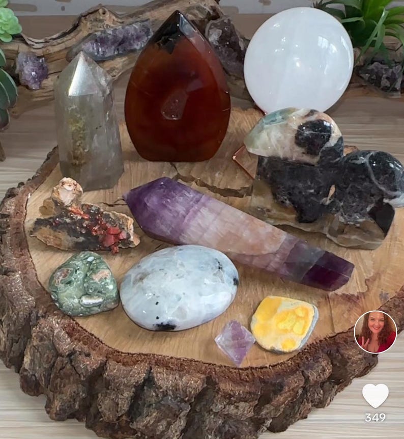 May include: A collection of crystals and stones on a wooden slice. The crystals include amethyst, clear quartz, carnelian, and a white selenite plate. The stones are green, brown, and black.