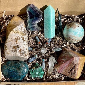 May include: A collection of crystals and stones in a box, including a green and blue crystal, a light blue crystal, a brown and white crystal, a green and blue sphere, a green and blue crystal cluster, a light blue crystal cluster, a brown and white crystal, a clear crystal, and a metal star-shaped object.
