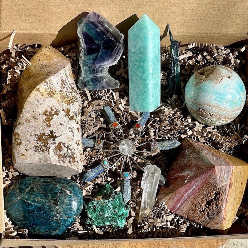 May include: A collection of crystals and stones in a box, including a green and blue crystal, a light blue crystal, a brown and white crystal, a green and blue sphere, a green and blue crystal cluster, a light blue crystal cluster, a brown and white crystal, a clear crystal, and a metal star-shaped object.