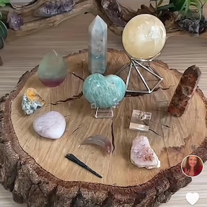 May include: A collection of crystals and stones arranged on a wooden slice. The crystals include a green heart-shaped crystal, a clear crystal point, a green and pink crystal, a clear crystal ball, a brown crystal, a white crystal, a pink crystal, and a gray crystal. The crystals are arranged in a circle around the wooden slice.