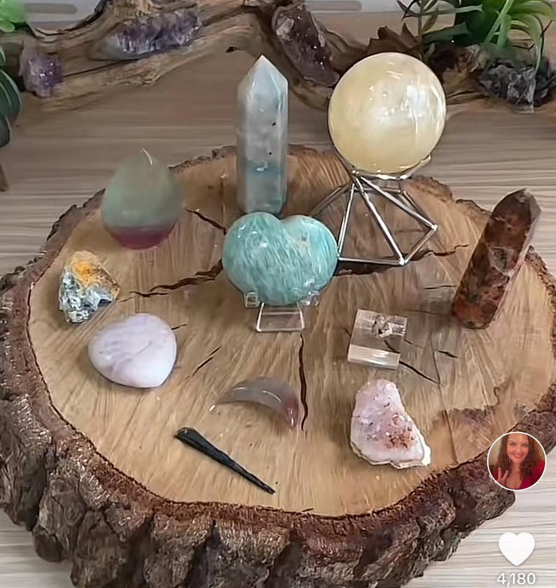 May include: A collection of crystals and stones arranged on a wooden slice. The crystals include a green heart-shaped crystal, a clear crystal point, a green and pink crystal, a clear crystal ball, a brown crystal, a white crystal, a pink crystal, and a gray crystal. The crystals are arranged in a circle around the wooden slice.