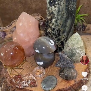 May include: A collection of polished stones and crystals arranged on a wooden slice. The stones include a large green and black crystal, a pink crystal, a gray and white agate, a brown sphere, and a green and white stone. The wooden slice is on a brown surface.