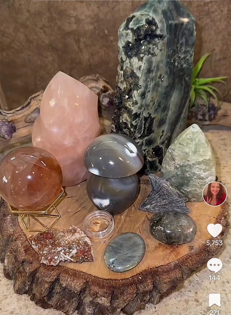 May include: A collection of polished stones and crystals arranged on a wooden slice. The stones include a large green and black crystal, a pink crystal, a gray and white agate, a brown sphere, and a green and white stone. The wooden slice is on a brown surface.