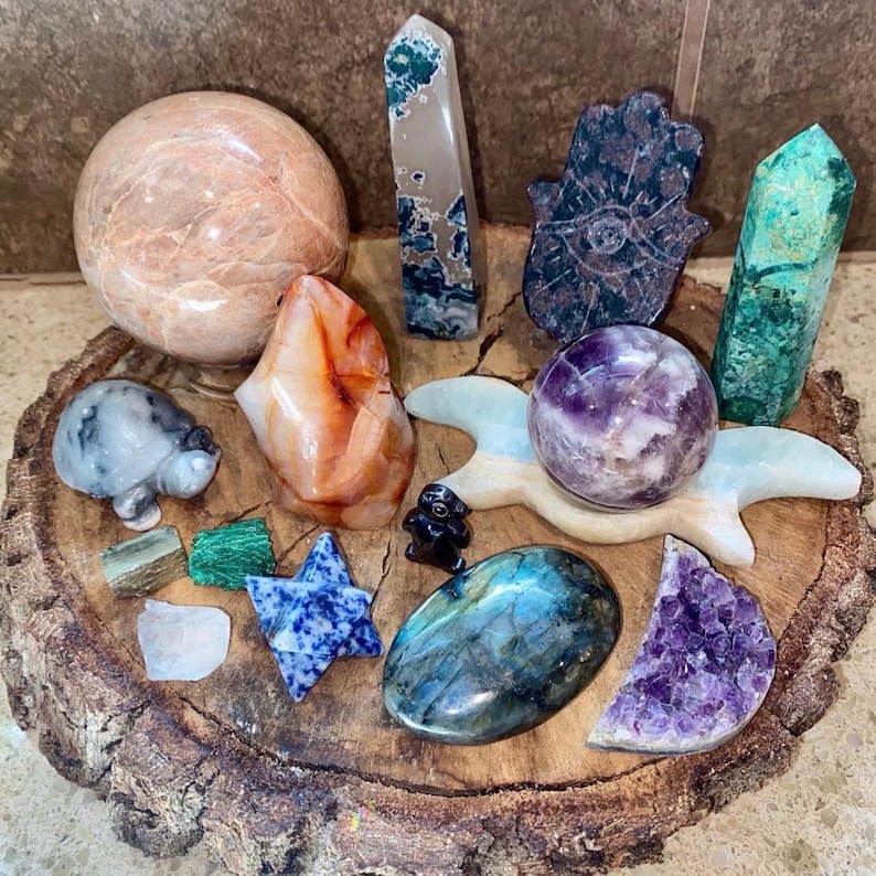 May include: A collection of polished crystals and stones, including a large orange carnelian, a green aventurine, a purple amethyst, a blue labradorite, a green jade, a pink rose quartz, a grey turtle, a blue star, a hamsa hand, and a white moon.