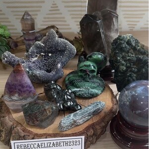 May include: A collection of crystals and stones, including amethyst, smoky quartz, green aventurine, and a crystal skull. The crystals are arranged on a wooden base and a sign reads "$555 Intuitive Crystal Set".