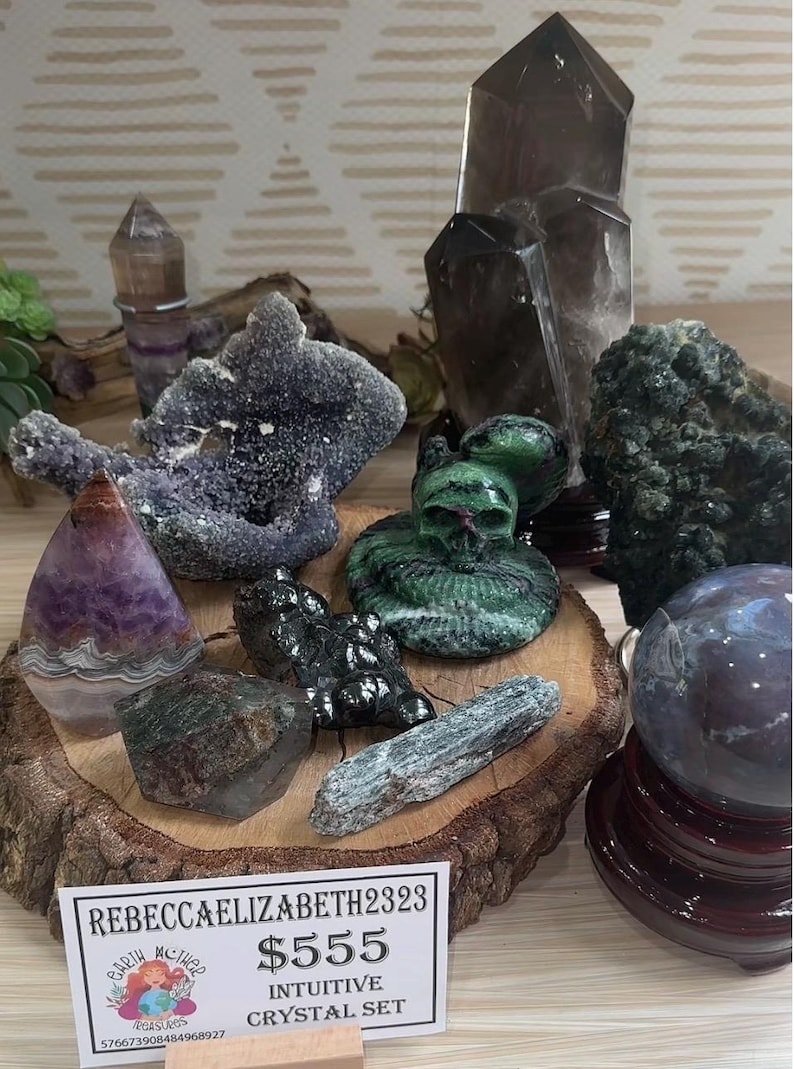 May include: A collection of crystals and stones, including amethyst, smoky quartz, green aventurine, and a crystal skull. The crystals are arranged on a wooden base and a sign reads "$555 Intuitive Crystal Set".