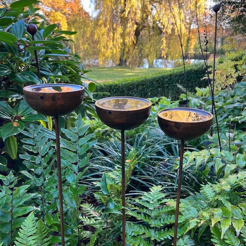 Large rain catchers made of metal which rust over time, filled with water, birds drawn to bathe, green foliage in background