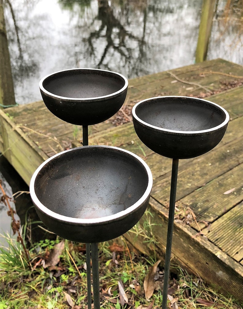 Large rain catchers made of metal which rust over time,  can be filled with water, birds drawn to bathe, jetty and pond in background