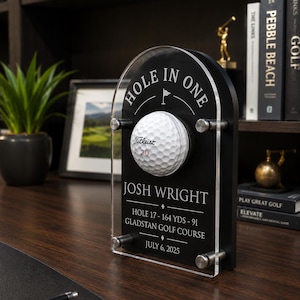 May include: A black arched acrylic display stand featuring a white Titleist 20 golf ball mounted in the centre. The stand is engraved with the text HOLE IN ONE, JOSH WRIGHT, HOLE 17 - 164 YDS - 91, GLADSTAN GOLF COURSE, and JULY 6, 2025.