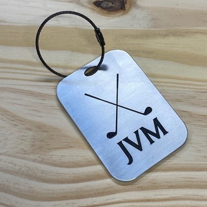 May include: Silver luggage tag with a black golf club design and the initials "JVM" engraved on it. The tag is attached to a black metal ring.
