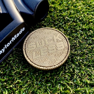 May include: A gold-colored golf ball marker with a geometric pattern and the words "SUCK LESS" embossed on the surface.